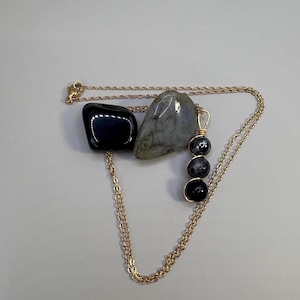 May include: A gold-colored chain necklace with a pendant of three black beads. The necklace is displayed with two stones: a black, polished stone and a gray, speckled stone. The background is a plain, light gray.