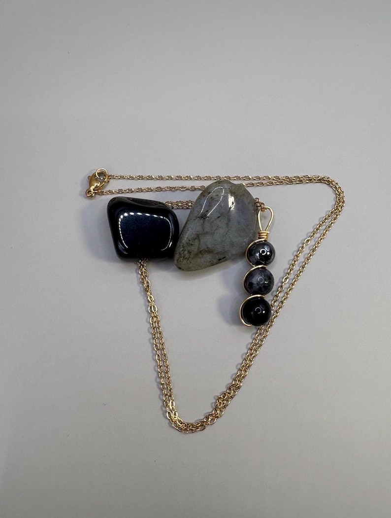 May include: A gold-colored chain necklace with a pendant of three black beads. The necklace is displayed with two stones: a black, polished stone and a gray, speckled stone. The background is a plain, light gray.