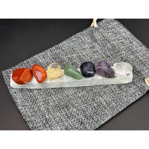 May include: Seven chakra stones, in the colors of the rainbow, are arranged on a selenite charging plate. The stones are red, orange, yellow, green, blue, indigo, and violet. The plate is white and has a rough texture.