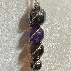 May include: A silver wire wrapped pendant necklace with four beads. The top and bottom beads are black hematite, the middle bead is amethyst, and the bead in between is smoky quartz.
