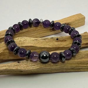 May include: A purple amethyst and black hematite beaded bracelet with a silver accent. The bracelet is on a wooden surface.