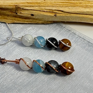 May include: Two necklaces with copper wire wrapped around a series of gemstone beads. The beads are white, blue, gray, and brown. The necklaces are on a light blue fabric background.