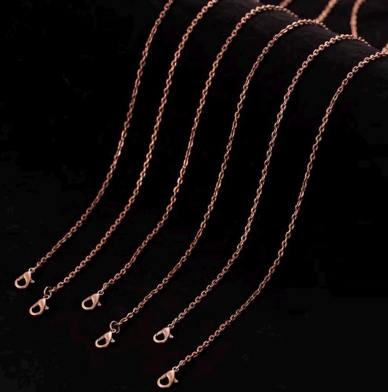 May include: Several rose gold-colored chain necklaces with lobster claw clasps are displayed against a dark background. The chains are thin and delicate, with a subtle shine, suggesting they are made of metal.