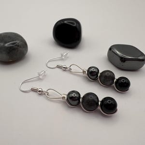 May include: A pair of dangle earrings featuring a silver wire design with three dark gray and black beads. The earrings are displayed with several polished stones in shades of gray and black, creating a minimalist aesthetic.