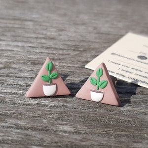 May include: A pair of triangle-shaped earrings with a green plant design in a white pot on a pink background.