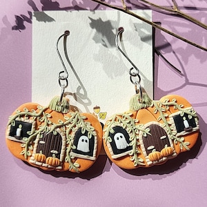 May include: Orange pumpkin-shaped earrings with a Halloween theme. Each earring features a house design with a ghost in the window, a door, and a candle. Green vines and small pumpkins decorate the earrings, which hang from silver hooks.