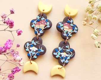 May include: A pair of dangle earrings with a woodland theme. Each earring features three brown clover-shaped charms with blue and white floral details, red mushrooms, and a yellow crescent moon charm.