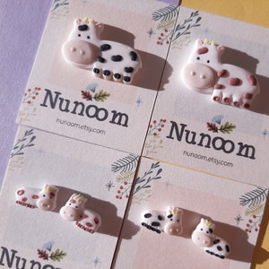 Pu&ograve; includere: Quattro paia di orecchini a forma di mucca e vitello. Gli orecchini sono realizzati in pasta polimerica e sono dipinti in rosa, bianco, nero e marrone. Gli orecchini sono esposti su cartoncini bianchi con il testo "Nunoom" e "nunoom.etsy.com".