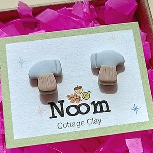 May include: A pair of small hammer-shaped earrings in a gift box. The earrings are made of gray and brown clay. The box is lined with pink tissue paper and has a white card with the text "Noom Cottage Clay".