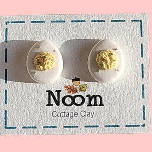 May include: A pair of white ceramic earrings shaped like deviled eggs. The earrings are on a white card with the text "Noom Cottage Clay".