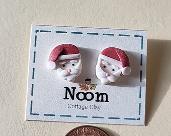 May include: A pair of red and white Santa Claus face earrings made of polymer clay. The earrings are displayed on a white card with the text "Noom Cottage Clay".