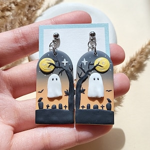 May include: A pair of Halloween-themed earrings featuring a ghost silhouette in front of a tree and a crescent moon. The earrings are made of polymer clay and have a black, orange, and yellow color scheme.