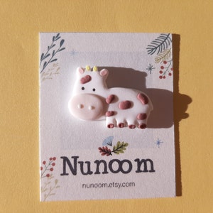 Pu&ograve; includere: Ippopotamo in ceramica bianca con macchie rosa e marroni. L'ippopotamo ha una corona gialla sulla testa. L'ippopotamo &egrave; su una carta bianca con un disegno floreale e il testo "Nunoom" e "nunoom.etsy.com".