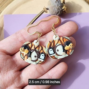 May include: A pair of gold dangle earrings with orange, green, and black pumpkin-shaped charms. The charms feature a witch's hat, a broom, and a ghost. The earrings are 2.5 cm / 0.98 inches long.