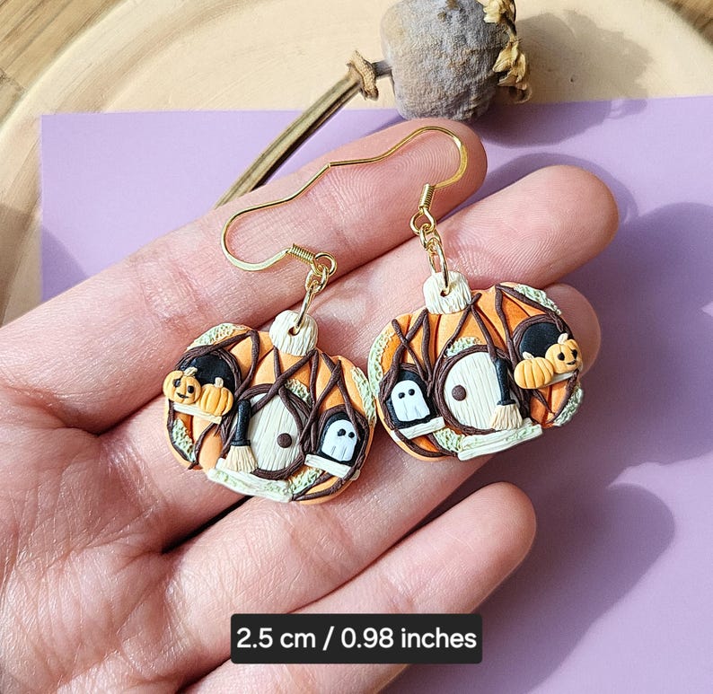 May include: A pair of gold dangle earrings with orange, green, and black pumpkin-shaped charms. The charms feature a witch's hat, a broom, and a ghost. The earrings are 2.5 cm / 0.98 inches long.