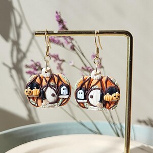 May include: A pair of orange pumpkin-shaped earrings with a small house, ghosts, and bats on each earring. The earrings are made of polymer clay and have gold hooks.