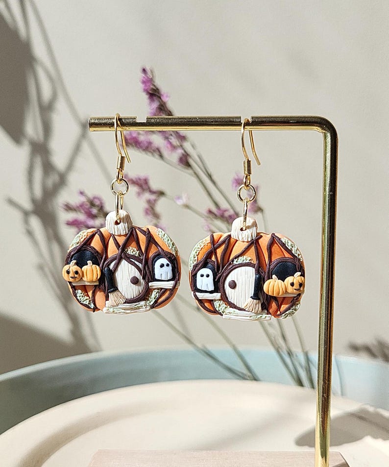 May include: A pair of orange pumpkin-shaped earrings with a small house, ghosts, and bats on each earring. The earrings are made of polymer clay and have gold hooks.