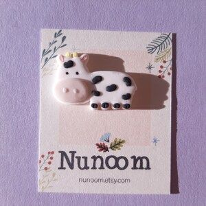 Pu&ograve; includere: Un ciondolo a forma di mucca rosa e nero con una stella gialla sulla testa. Il ciondolo &egrave; su una carta bianca con il testo "Nunoom" e "nunoom.etsy.com".