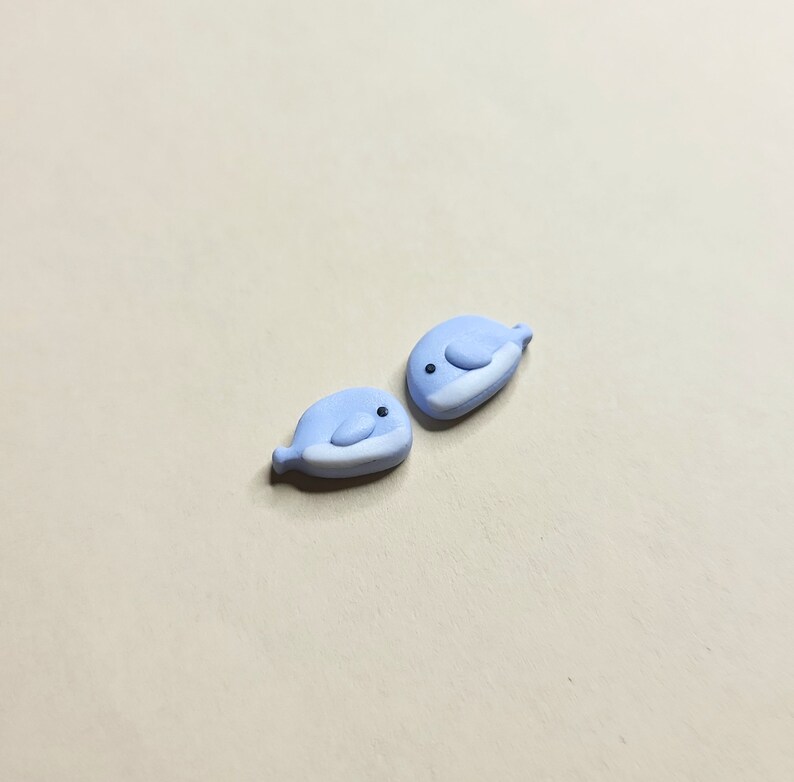 May include: Two small, light blue whale figurines with white bellies and black eyes. The whales are facing each other.