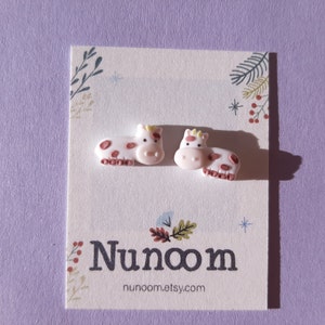 Pu&ograve; includere: Un paio di orecchini a bottone a forma di mucca rosa con macchie marroni. Gli orecchini sono su una carta bianca con il testo "Nunoom" e "nunoom.etsy.com".