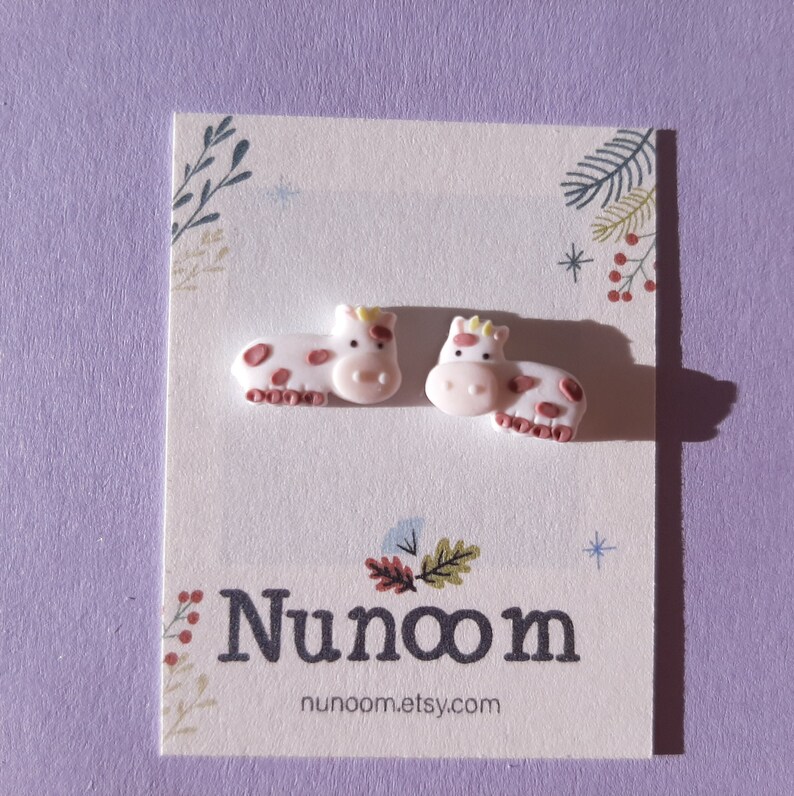 Pu&ograve; includere: Un paio di orecchini a bottone a forma di mucca rosa con macchie marroni. Gli orecchini sono su una carta bianca con il testo "Nunoom" e "nunoom.etsy.com".