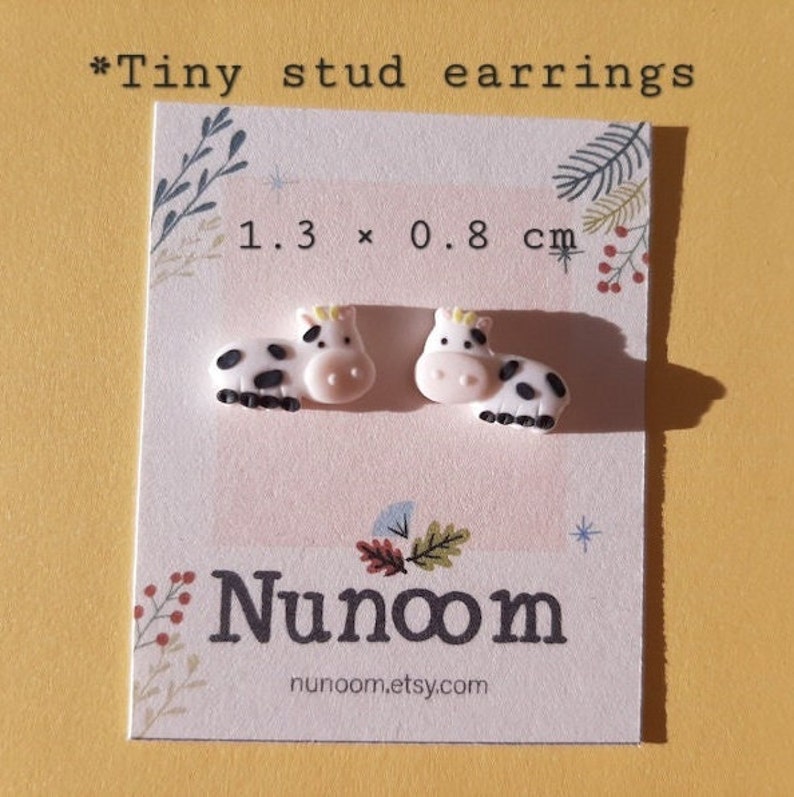 Pu&ograve; includere: Un paio di piccoli orecchini a forma di mucca su una carta bianca con il testo "1,3 x 0,8 cm" e "Nunoom" con l'indirizzo del sito web "nunoom.etsy.com". Gli orecchini sono bianchi con macchie nere e dettagli gialli.