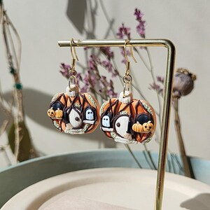 May include: A pair of orange and brown Halloween-themed earrings. Each earring features a pumpkin with a small house, a ghost, and a witch's hat.