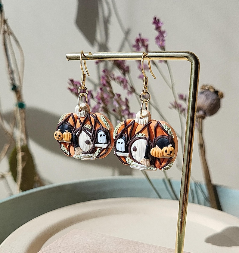 May include: A pair of orange and brown Halloween-themed earrings. Each earring features a pumpkin with a small house, a ghost, and a witch's hat.