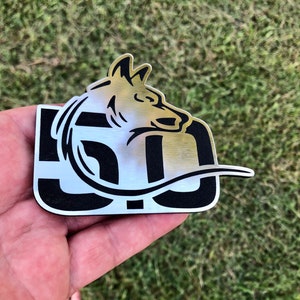 May include: A silver metal emblem with a black outline of a wolf's head and a wave. The wolf's head is facing the left and the wave is below the wolf's head. The emblem is on a hand.