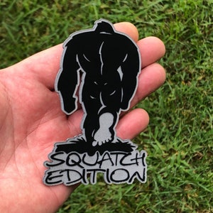 May include: A silver-colored patch with a black silhouette of Bigfoot walking away from the viewer. The text "SQUATCH EDITION" is printed below the silhouette.