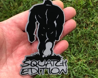 May include: A silver-colored patch with a black silhouette of Bigfoot walking away from the viewer. The text "SQUATCH EDITION" is printed below the silhouette.