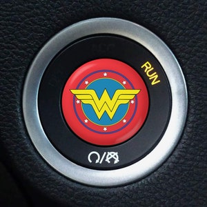May include: A black car ignition button with a silver trim. The button features the Wonder Woman logo in the center, with a red background, blue center, and yellow logo. The word "RUN" is printed in yellow.