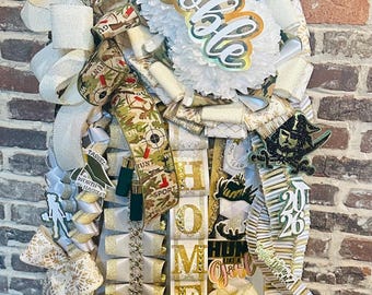 Puede incluir: Una gran decoración de tipo homecoming mum con cintas blancas, doradas y de camuflaje. Incluye una flor blanca central con el nombre Noble, un letrero de Class of 2026 y letras doradas verticales que forman HOMECOMING. Varios detalles incluyen un casco de fútbol americano, un lazo pequeño y gráficos con temática de caza.