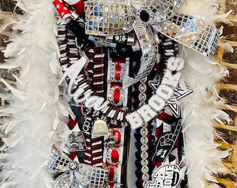 Puede incluir: Un homecoming mum con una boa de plumas blancas, cintas superpuestas en rojo, negro, blanco y plata, y un gran lazo de bola de discoteca plateado. Incluye letras brillantes que dicen "Ava Brooks" y "HOMECOMING 2025" con un gráfico de casco.
