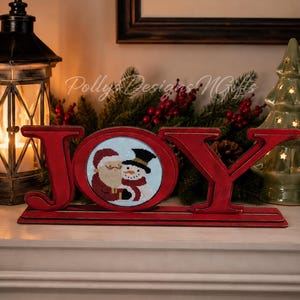 May include: A red wooden "JOY" sign with a Santa and snowman cross-stitch design in the "O". The sign is on a white mantel with a lantern and Christmas tree in the background. The sign is a festive holiday decoration.
