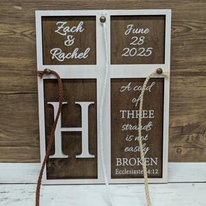 May include: A wooden wedding sign with a white frame and brown panels. The sign features the names "Zach & Rachel", the date "June 28 2025", the letter "H", and the quote "A cord of three strands is not easily BROKEN". Braided cords hang from the top.
