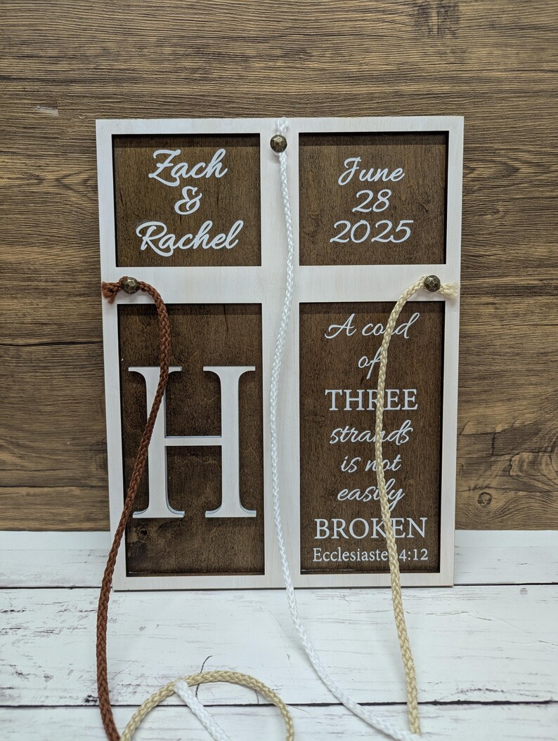 May include: A wooden wedding sign with a white frame and brown panels. The sign features the names "Zach & Rachel", the date "June 28 2025", the letter "H", and the quote "A cord of three strands is not easily BROKEN". Braided cords hang from the top.