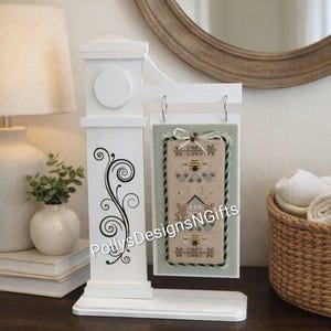 May include: A white decorative sign holder with a black swirl design on the side. The sign holder displays a framed cross-stitch with bees and the text "PollysDesignsNGifts". The sign is hanging from metal rings.