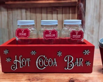 May include: Red wooden box with the words "Hot Cocoa Bar" on the front. Three glass jars with white lids and labels are inside the box. The labels read "Cocoa Mix", "Marshmallows", and "Peppermint".