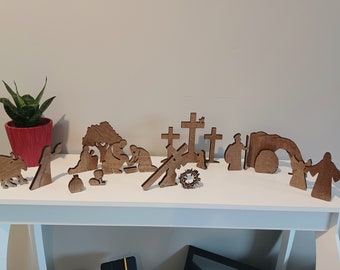 May include: A set of wooden laser cut figures depicting the story of Easter. The figures include a garden, a cross, a tomb, and figures of people. The figures are all brown and are arranged on a white shelf.