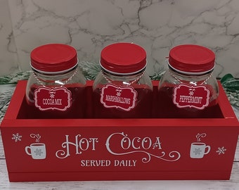 May include: Red wooden box with three glass jars labeled "Cocoa Mix", "Marshmallows", and "Peppermint". The box has a white design that says "Hot Cocoa Served Daily".