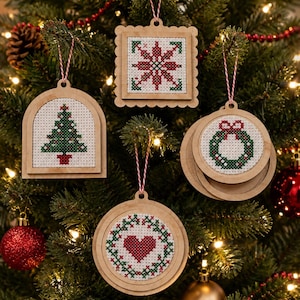 May include: Four wooden ornaments featuring cross-stitch designs on white fabric. The shapes include an arch with a Christmas tree, a square with a star, and two circles featuring a wreath and a heart. Each ornament hangs from a red and white string.