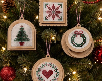 May include: Four wooden ornaments featuring cross-stitch designs on white fabric. The shapes include an arch with a Christmas tree, a square with a star, and two circles featuring a wreath and a heart. Each ornament hangs from a red and white string.