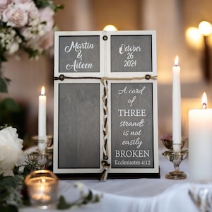 May include: A wooden wedding sign with a white frame and gray background. The sign displays the names "Martin & Aileen" and the date "October 26 2024". It also features the text "A cord of THREE strands is not easily BROKEN".