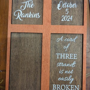 May include: Wooden sign with an orange frame and four sections. The top left section reads "The Rankins" in white script. The top right section says "October 5 2024". The bottom right section has the text "A cord of THREE strands is not easily BROKEN".