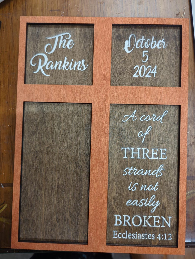 May include: Wooden sign with an orange frame and four sections. The top left section reads "The Rankins" in white script. The top right section says "October 5 2024". The bottom right section has the text "A cord of THREE strands is not easily BROKEN".
