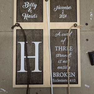 May include: Wooden sign with a light wood frame and dark brown interior panels. The sign features the names "Billy & Heidi", the date "November 2 2024", the letter "H", and the quote "A cord of THREE strands is not easily BROKEN".