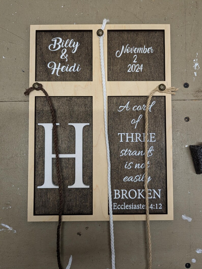 May include: Wooden sign with a light wood frame and dark brown interior panels. The sign features the names "Billy & Heidi", the date "November 2 2024", the letter "H", and the quote "A cord of THREE strands is not easily BROKEN".