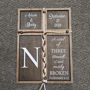 May include: Wooden framed sign with four sections. The top left section reads "Adam & Becky", the top right "September 21 2024". The bottom left has a large "N", and the bottom right reads "A cord of THREE strands is not easily BROKEN Ecclesiastes 4:12".