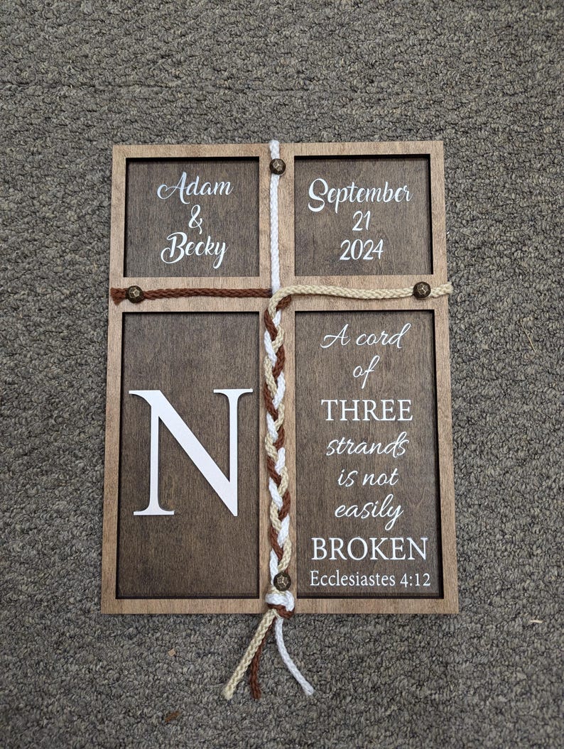 May include: Wooden framed sign with four sections. The top left section reads "Adam & Becky", the top right "September 21 2024". The bottom left has a large "N", and the bottom right reads "A cord of THREE strands is not easily BROKEN Ecclesiastes 4:12".